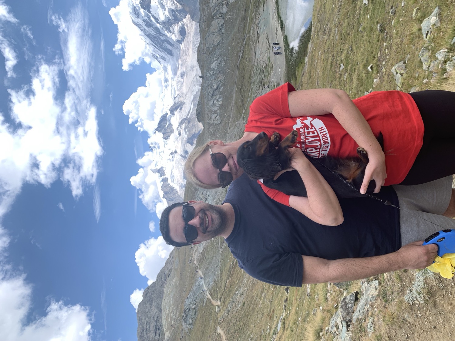 Jirina and Adam holding Aja, their miniature Dachshund, on a mountain path in the Swiss Alps with a glacier behind them