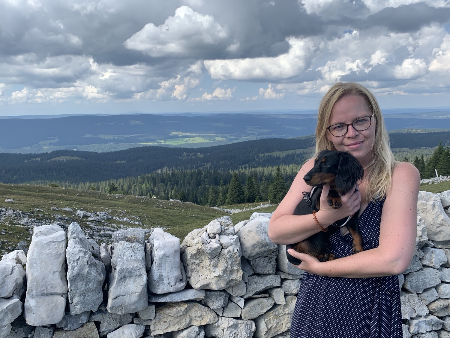 Jirina holding Aja beside a dry stone wall on a Jura ridge, with forested hills stretching behind
